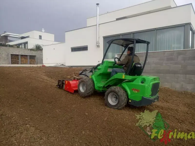 Profesionálne terénne úpravy pozemku pomocou zeleného mini nakladača s červeným príslušenstvom pri modernom dome s bielou fasádou